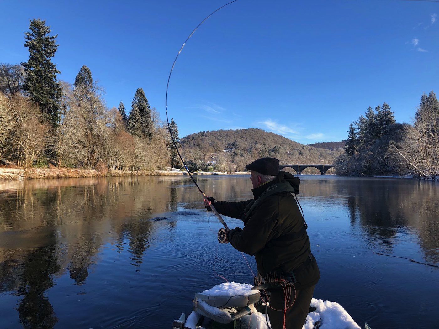 This River Tay fisher is Spey casting from the traditional Tay salmon fishing boat during late January on the Tay at Dunkeld. If you look closely you'll see the 200 year old Telford Bridge in the distance which is one of the Tay's most historic & beautiful sandstone bridges. You can see this fly fisher is wrapped up warmly due to the early Spring cold weather conditions.